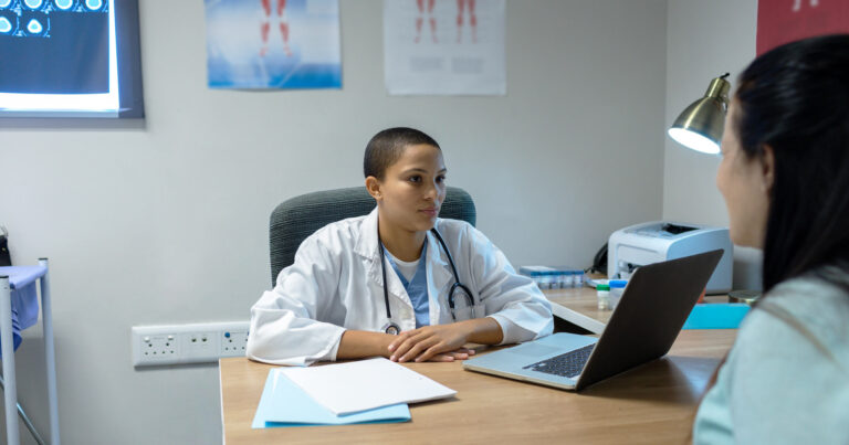 Un médecin regarde un patient au-dessus de son bureau, tandis qu’un patient explique sa situation.