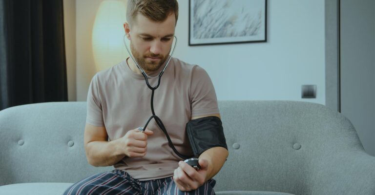 A man sits on the couch in his pajamas and gives himself a blood pressure check.