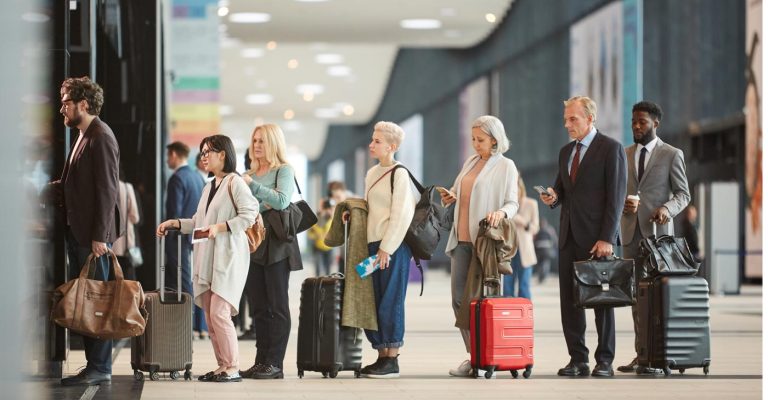 Les voyageurs avec des valises s’alignent à un kiosque de l’aéroport, regardant leur téléphone et ayant l’air fatigué.