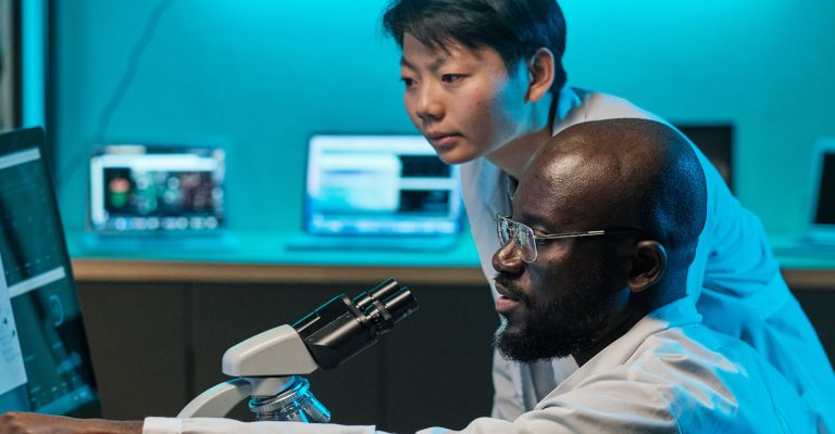 Deux chercheurs se tiennent au-dessus d’un bureau avec un microscope et des écrans d’ordinateur, discutant de quelque chose de difficile.