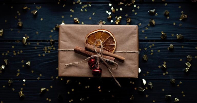 Un cadeau de Noël, élégamment enveloppé dans une tranche d’orange et une ficelle brune, trône sur une table sombre recouverte d’étoiles dorées.