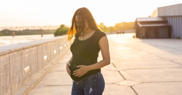 A woman stands in a courtyard, pregnant, happy in the glowing light of the late afternoon.