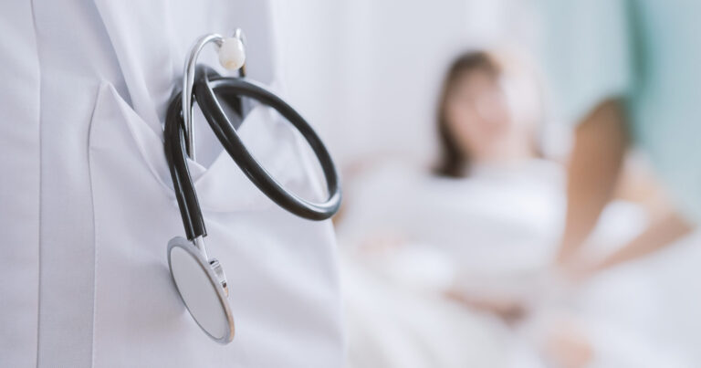 Closeup of a stethoscope hanging out of a doctor's coat pocket with a background of a patient lying in a bed, but out of focus. 