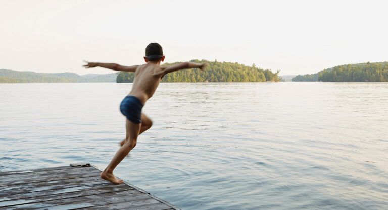 A young person gleefully runs and jumps off a dock into a stunning lake at a summer camp.